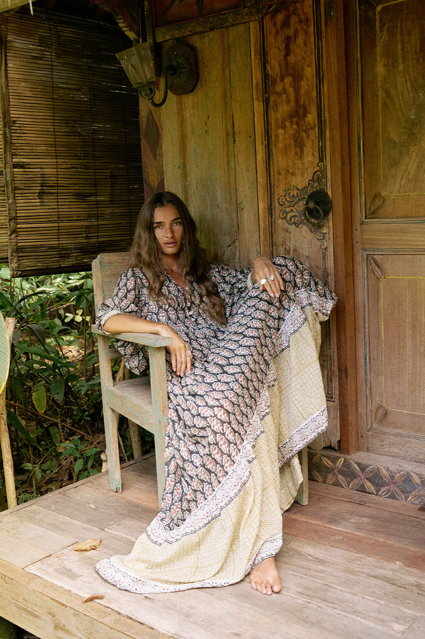 Bhoomi Maxi Dress Onyx — full length seated on rustic wooden veranda leaning on chair, onyx hand block print maxi dress with tiered hem, old wooden doors and bamboo wall | Daughters of India