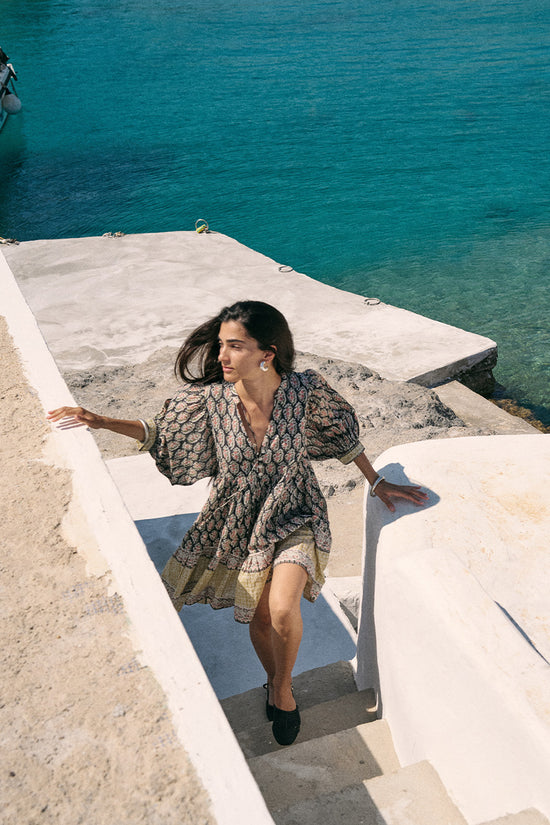 Woman on a seaside stone ledge overlooking turquoise water wearing the Daughters of India Bhoomi Mini Dress in Onyx, the dark block print fabric contrasting with the bright sea