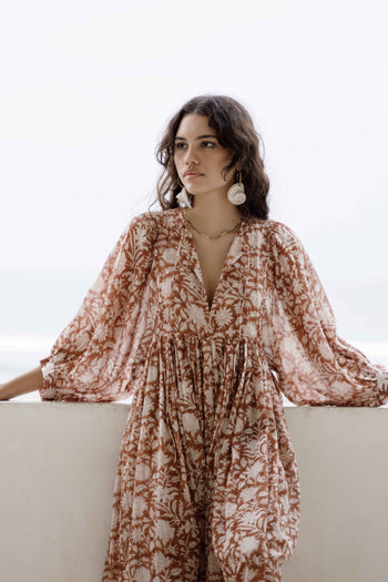 Daughters of India Kyra Midi Dress in Caramel — upper body leaning on veranda wall, V-neckline, billowed sleeves