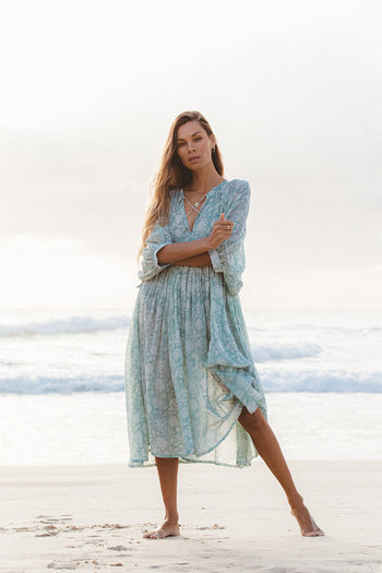 Woman wearing Daughters of India Kyra Midi Dress in Jade, a floral blue-green chiffon dress, standing on a beach