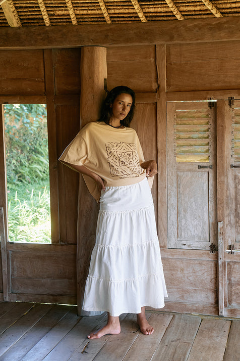 Full-length view of woman wearing the Daughters of India Shanti Tee in Cashew with a white tiered maxi skirt in a rustic wooden interior