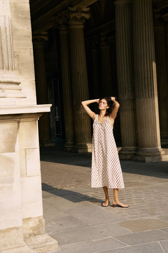 Woman standing between classical columns in Paris wearing the Daughters of India Sundress Midi in Peony, full-length view with flat sandals