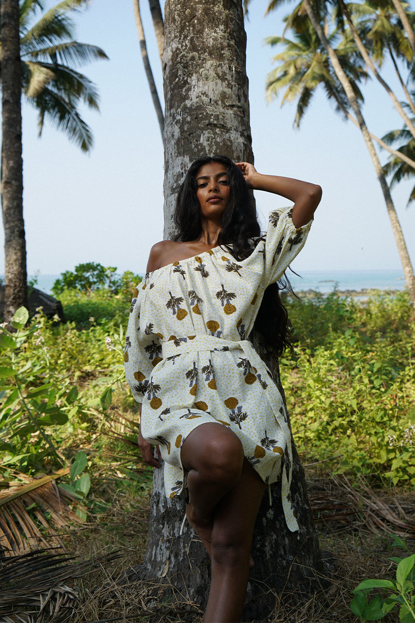 Dahlia Mini Dress Marigold — model leaning against palm tree, tropical beach backdrop, off-shoulder with tied waist, white linen marigold hand block print mini, boho resort beach dress | Daughters of India